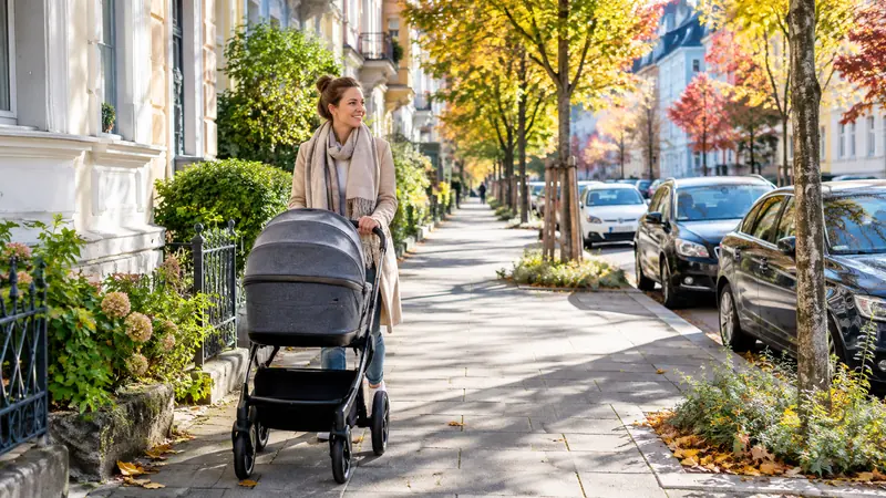 Mutter schiebt einen Kinderwagen über einen freien Gehweg, die Autos parken am Fahrbahnrand