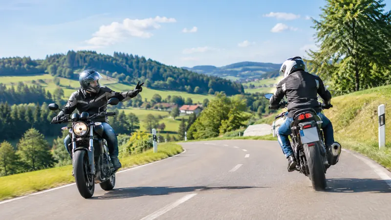 Zwei Motorradfahrer begegnen sich auf einer Landstraße, einer grüßt mit zwei Fingern